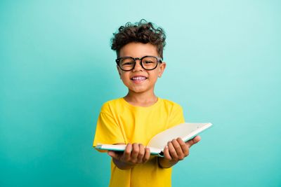 Happy boy in glasses holding an open book against a turquoise background.