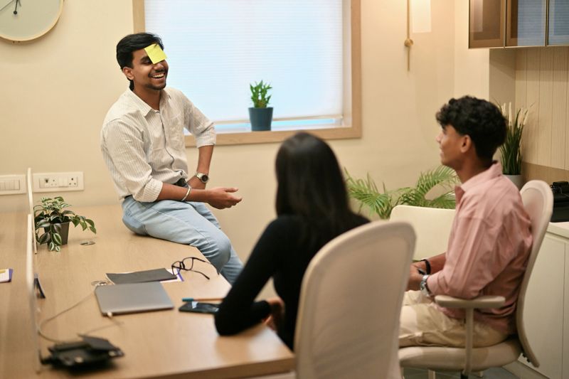 A young office worker with a sticky note on his forehead plays a fun game with two colleagues, capturing a joyful break in a relaxed, modern workplace.