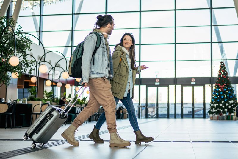 Young couple walking through airport terminal pulling suitcase and wearing backpacks during christmas season