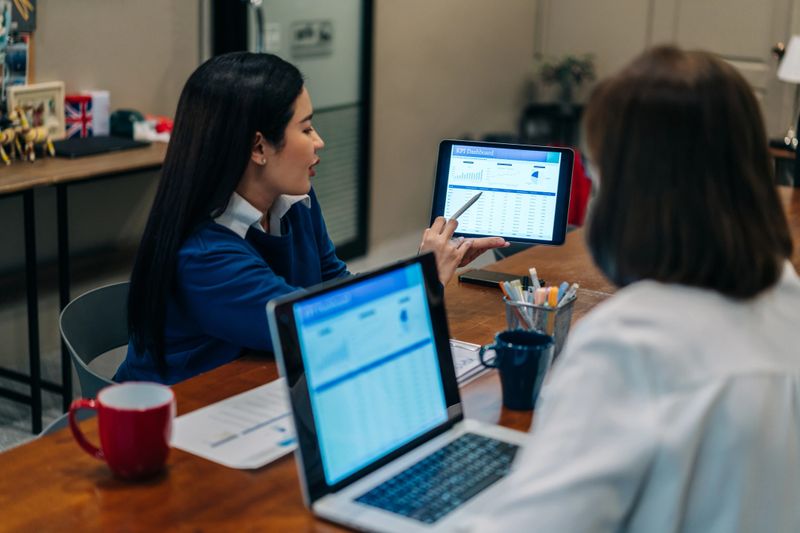 In a modern office, a young woman points to a KPI dashboard displayed on a digital tablet while speaking with a colleague seated beside her. Both professionals are engaged in a data-driven discussion, with a laptop, paperwork, and coffee mugs on the table, indicating a collaborative meeting. The image captures a focused, analytical moment in a workplace that combines digital tools with teamwork, likely centered around performance evaluation, marketing, or business intelligence in a casual but productive environment.