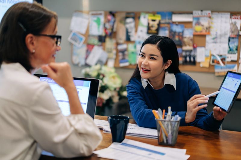 In a modern office, a young woman points to a KPI dashboard displayed on a digital tablet while speaking with a colleague seated beside her. Both professionals are engaged in a data-driven discussion, with a laptop, paperwork, and coffee mugs on the table, indicating a collaborative meeting. The image captures a focused, analytical moment in a workplace that combines digital tools with teamwork, likely centered around performance evaluation, marketing, or business intelligence in a casual but productive environment.