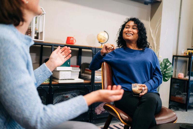 Two professional women are seated in a cozy, modern office lounge, engaged in a friendly and relaxed conversation. One woman sits on a leather sofa, while the other is in a chair across a coffee table, both holding drinks. The surrounding decor includes books, a globe, indoor plants, and a miniature greenhouse model, creating a warm, personalized workspace. The image reflects informal communication, workplace connection, and emotional intelligence in a creative, inclusive business environment.