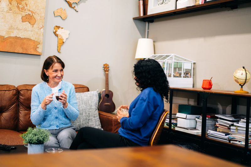 Two professional women are seated in a cozy, modern office lounge, engaged in a friendly and relaxed conversation. One woman sits on a leather sofa, while the other is in a chair across a coffee table, both holding drinks. The surrounding decor includes books, a globe, indoor plants, and a miniature greenhouse model, creating a warm, personalized workspace. The image reflects informal communication, workplace connection, and emotional intelligence in a creative, inclusive business environment.