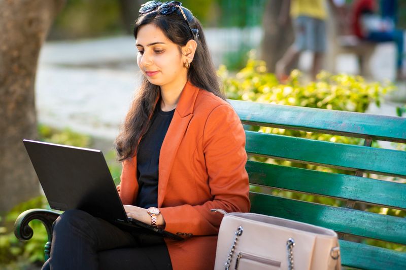 Portrait of happy Successful attractive indian young businesswoman in formal wear with sunglasses on her head sitting relaxed on a park bench with purse  and working outside on her laptop about the company in the park at day time. Young professional woman working on laptop and engaged in a productive work session.