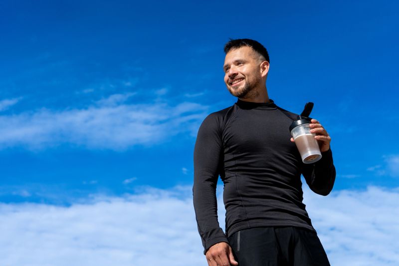 Fit man wearing black sportswear holding protein drink smiling confidently under bright blue sky after outdoor workout, healthy and active lifestyle.