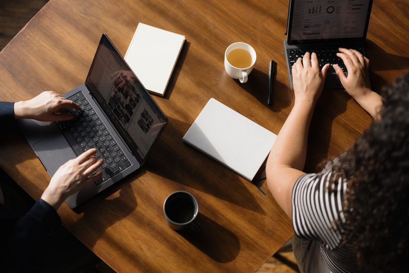 Two coworkers are working together on their laptops at a wooden table, enjoying coffee and tea
