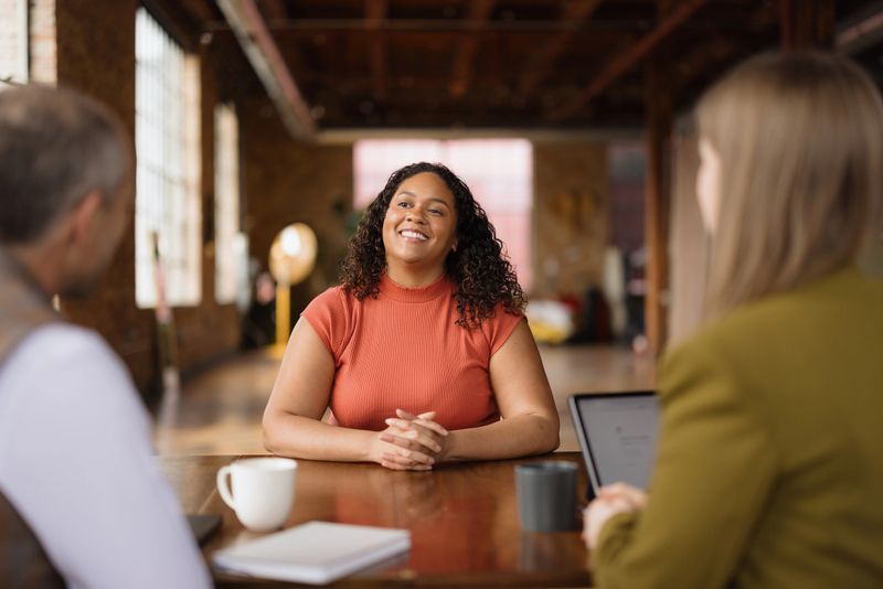 Smiling young woman sitting at a table with her hands clasped, attending a job interview with two recruiters