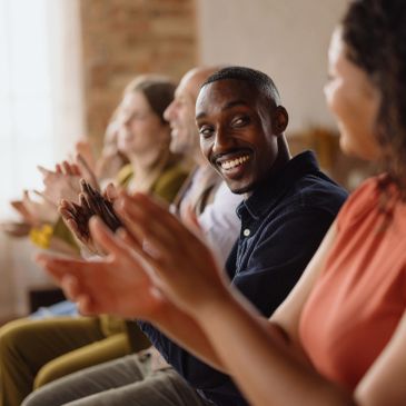 A group of diverse people clapping and smiling in a warm, casual setting.