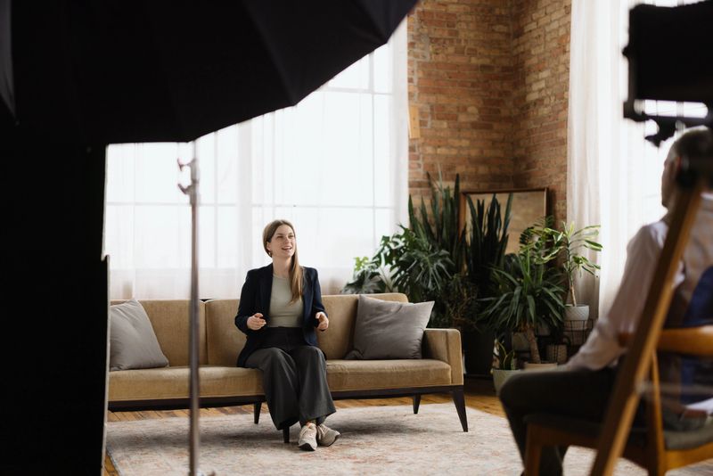 Young woman sitting comfortably on a sofa, engaging in an interview with a journalist in a stylish loft apartment, surrounded by modern decor