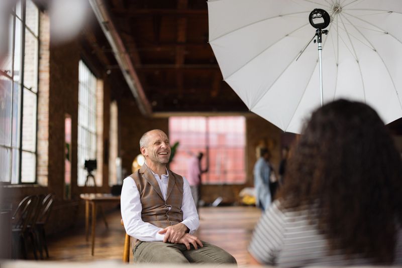 Cheerful mature businessman sitting in a modern loft, engaging in an interview, laughing and sharing insights with a photographer