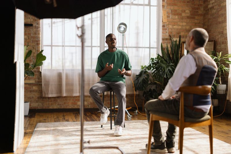 Young man sitting on a stool, engaging in an interview with a journalist in a stylish loft, surrounded by natural light and a casual atmosphere