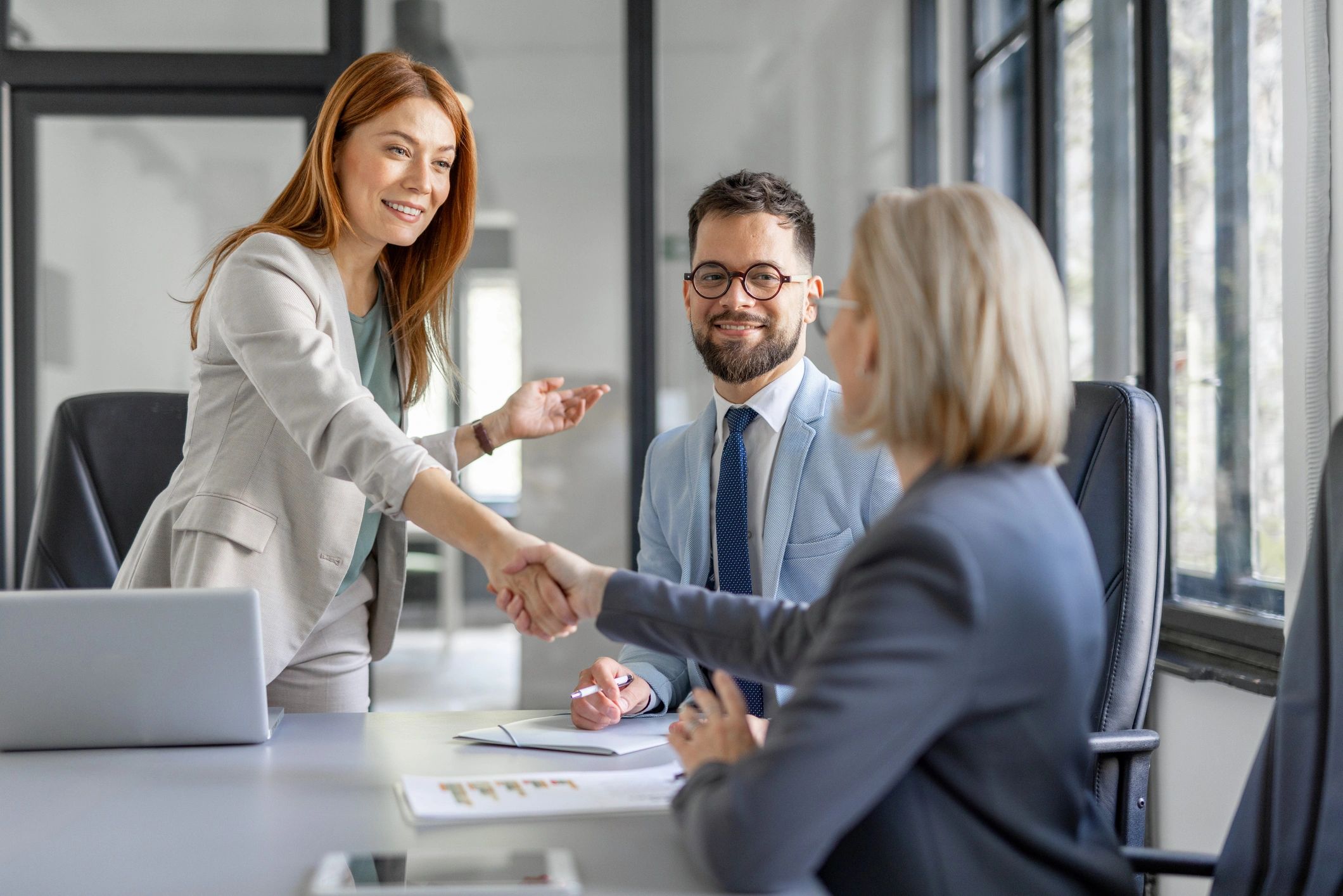 Two businesswomen shake hands in a modern office, while a businessman watches with a smile.