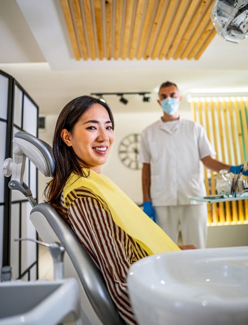Cheerful young woman smiles while sitting on a dentist chair, ready for her dental checkup, with the dentist in the background