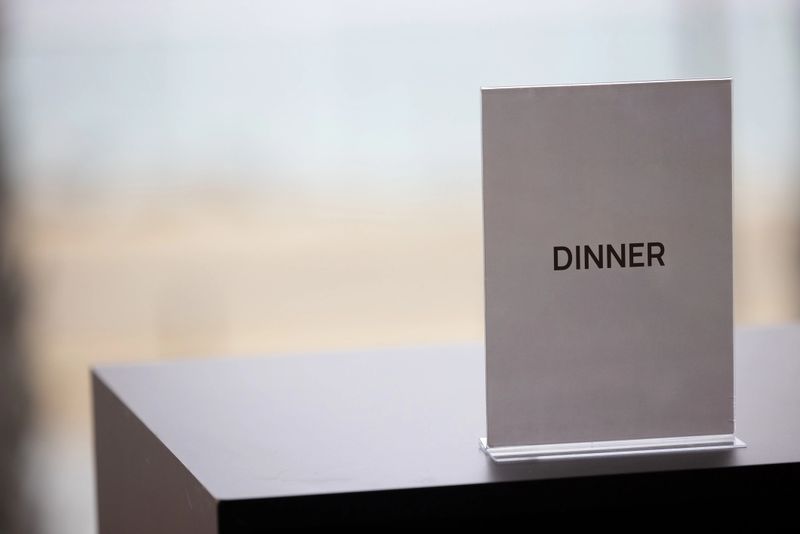 Minimalist acrylic tabletop sign in an elegant corporate setting. Blank white display card placed on a sleek wooden surface, with soft natural light from a blurred window background. Clean, professional environment ideal for business, hospitality, or event contexts. Displaying the sign “Dinner” for travel or event organization.