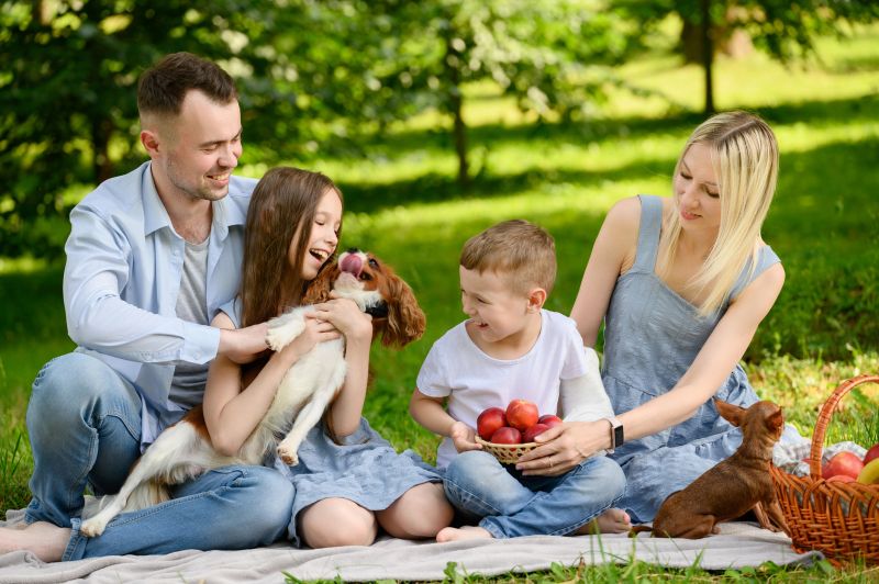 Joyful family enjoying picnic in park, having fun with playful dog, laughing happily. Mom, dad, brother and sister enjoying communication sitting on lawn. Family weekend concept.