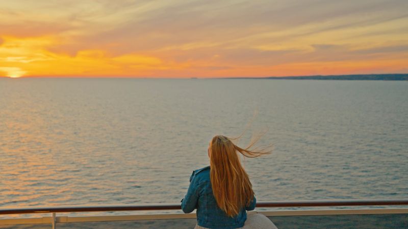 A mid adult Caucasian female is standing on a cruise ship deck, admiring a breathtaking sunset over the ocean. Her hair is gently blowing in the breeze, creating a serene and contemplative atmosphere.