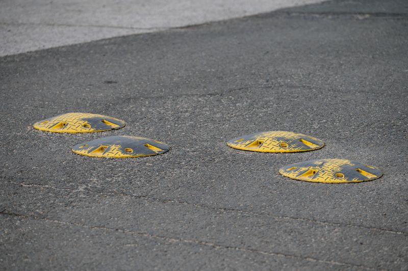 Four yellow speed bumps are embedded in a textured asphalt road, captured from a close angle in daylight. The distinct color and shape of the bumps emphasize their function in traffic calming and pedestrian safety within an urban or suburban setting.