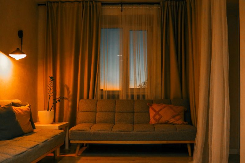 Cozy living room interior with warm lighting, grey sofas, decorative pillows, and a potted plant near the window during sunset.