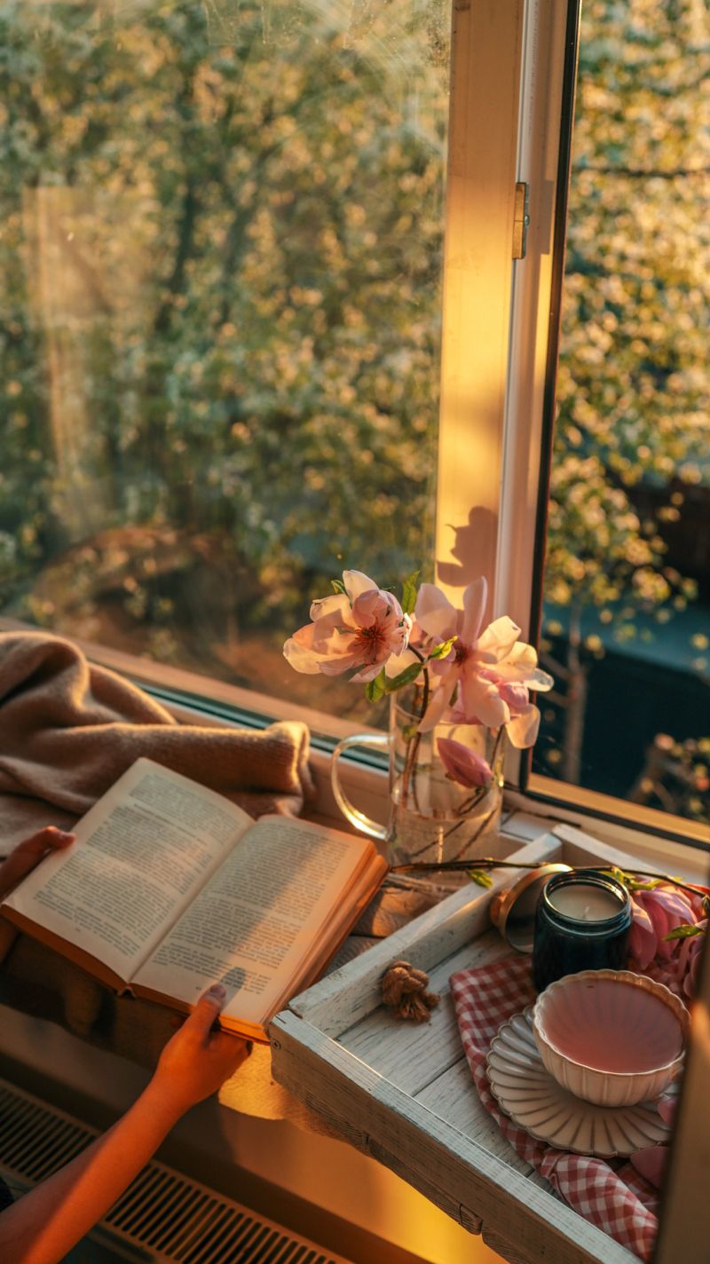 Person reading a book by a cozy window at sunset, with magnolia flowers, a black candle, and a cup of tea on a wooden tray. Warm golden light creates a peaceful, serene mood.