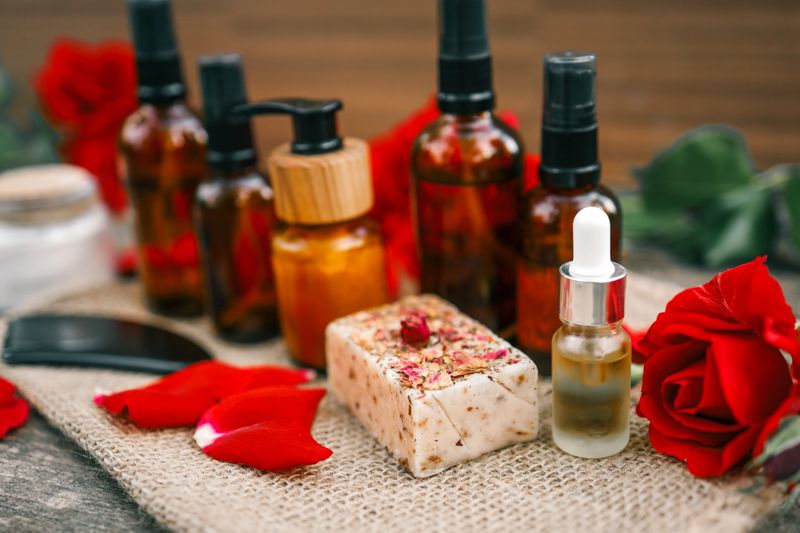 Handcrafted rose soap bar with dried petals, essential oil dropper and amber bottles arranged on a rustic burlap cloth with fresh rose petals.