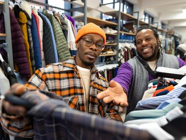 Two men shopping for clothes in a store, one showing a plaid shirt.