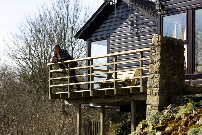 A mid-adult male stands on a wooden balcony, surrounded by leafless trees under a clear blue sky. Dressed casually in a flannel shirt and a t-shirt, he gazes into the distance, holding a cup of tea.  Located in Hexham, Northumberland.Videos of this scenario are available.