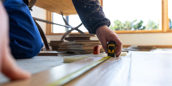Person measuring wooden floorboards with a tape measure indoors.