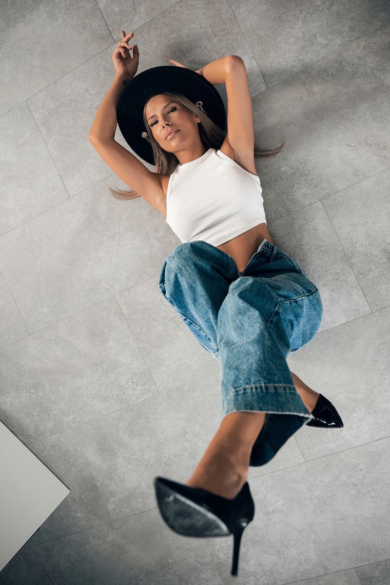 A woman in a crop top and jeans lounges on a light-colored floor while wearing a black hat and heels.
