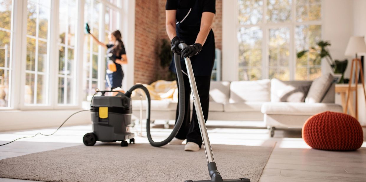 Two people cleaning a bright living room, vacuuming and wiping windows.