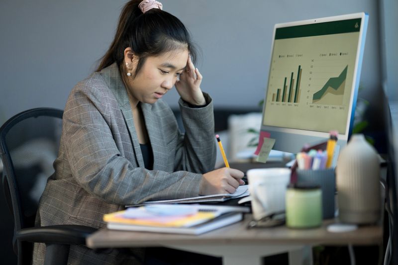 A professional woman focuses on analyzing data charts on her desktop computer, working in a modern office setting to interpret and record key business insights.