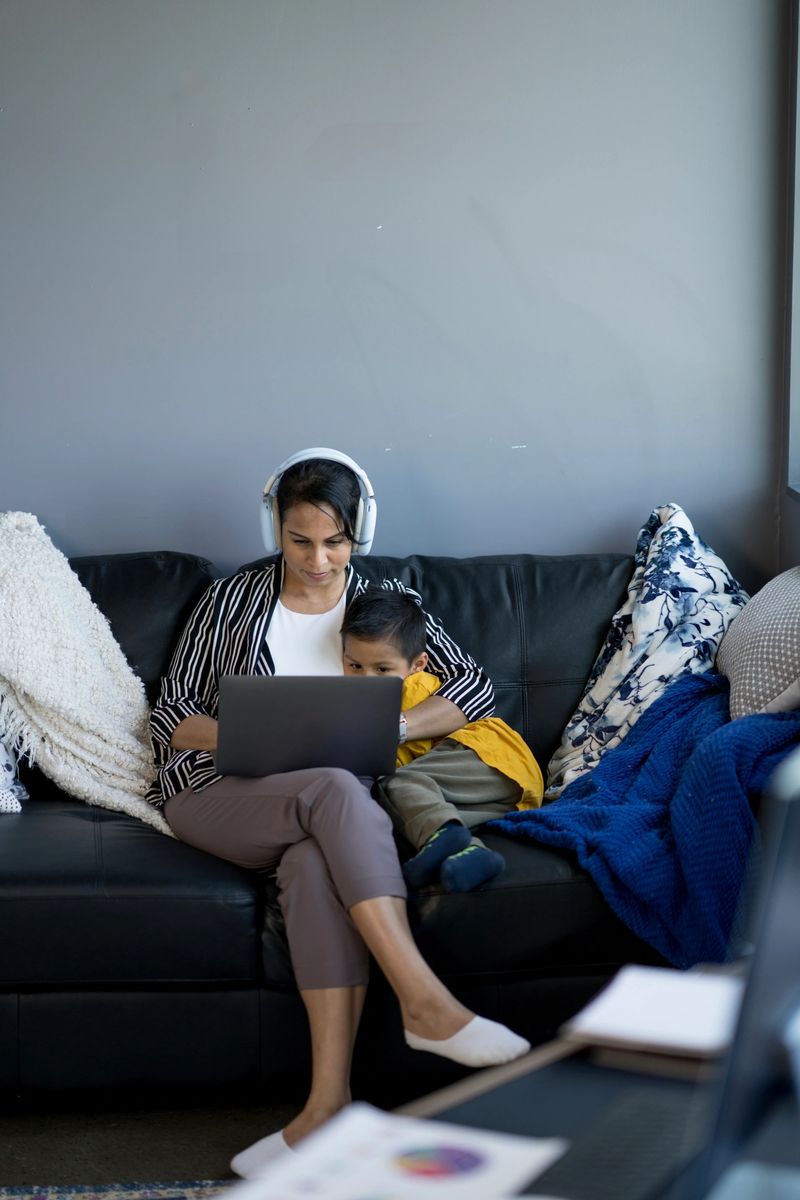 A mother wearing headphones and using a laptop works remotely while her son sits beside her on a sofa. The setting is a pleasant, modern living room, showcasing family time and multitasking dynamics.