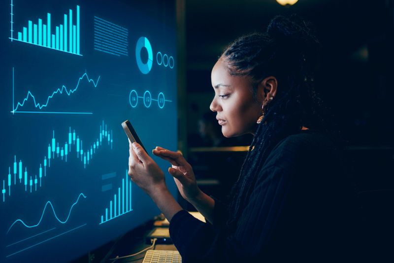 A professional Black woman analyzes data on her smartphone, illuminated by glowing graphs and charts on a large digital screen in a modern workspace.