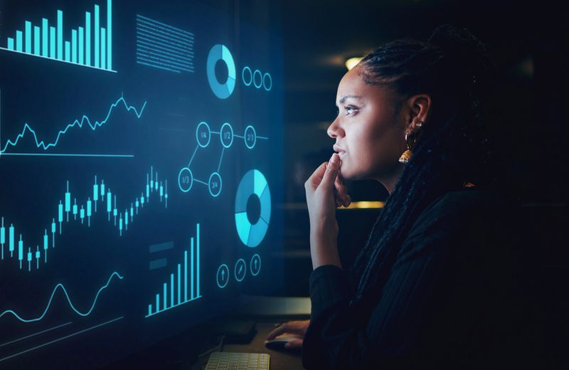 A thoughtful young woman with dark braided hair analyzes data on a glowing blue screen featuring charts, graphs, and metrics in a dimly lit workspace at night.