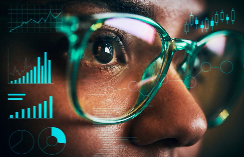 Close-up of a young woman wearing teal glasses, her focused gaze reflecting data analysis. Futuristic charts and graphs overlay the image, symbolizing modern technology in business or science.