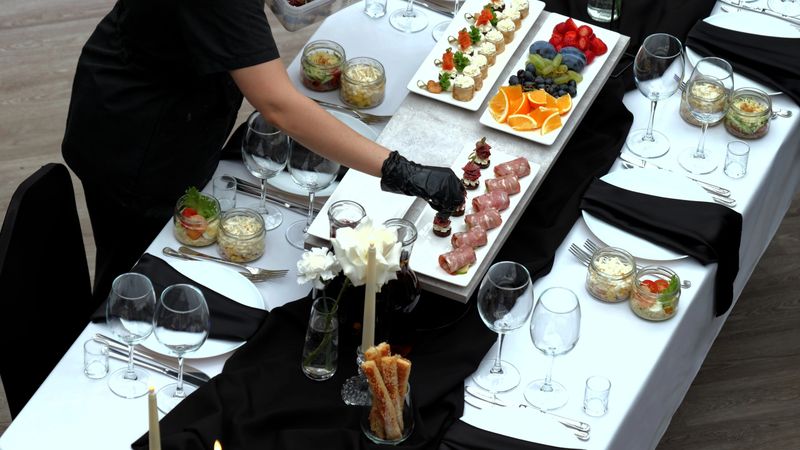Waiter wearing black gloves carefully placing gourmet appetizers on crisp white tablecloth, surrounded by elegant black napkins and softly glowing candles during upscale dining event