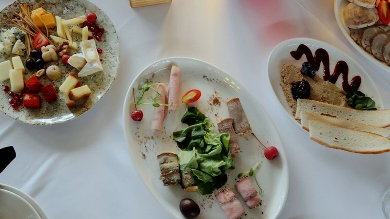 Variety of gourmet food, including cheese, cold cuts, pate, fruits and bread, served on white plates during a catered event, creating a sophisticated and celebratory atmosphere