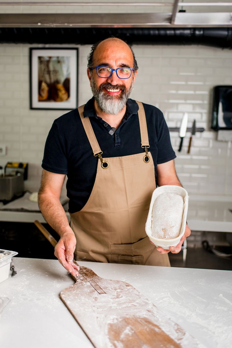 A close-up view of a person recording a bearded male baker  in a professional kitchen. The baker, wearing an apron, is standing behind a table full of various artisanal bread loaves. The image captures a modern content creation scene where the chef appears in focus, while the real-life background is blurred. Ideal for concepts related to food blogging, digital content creation, bakery business, small business marketing, and social media video production.