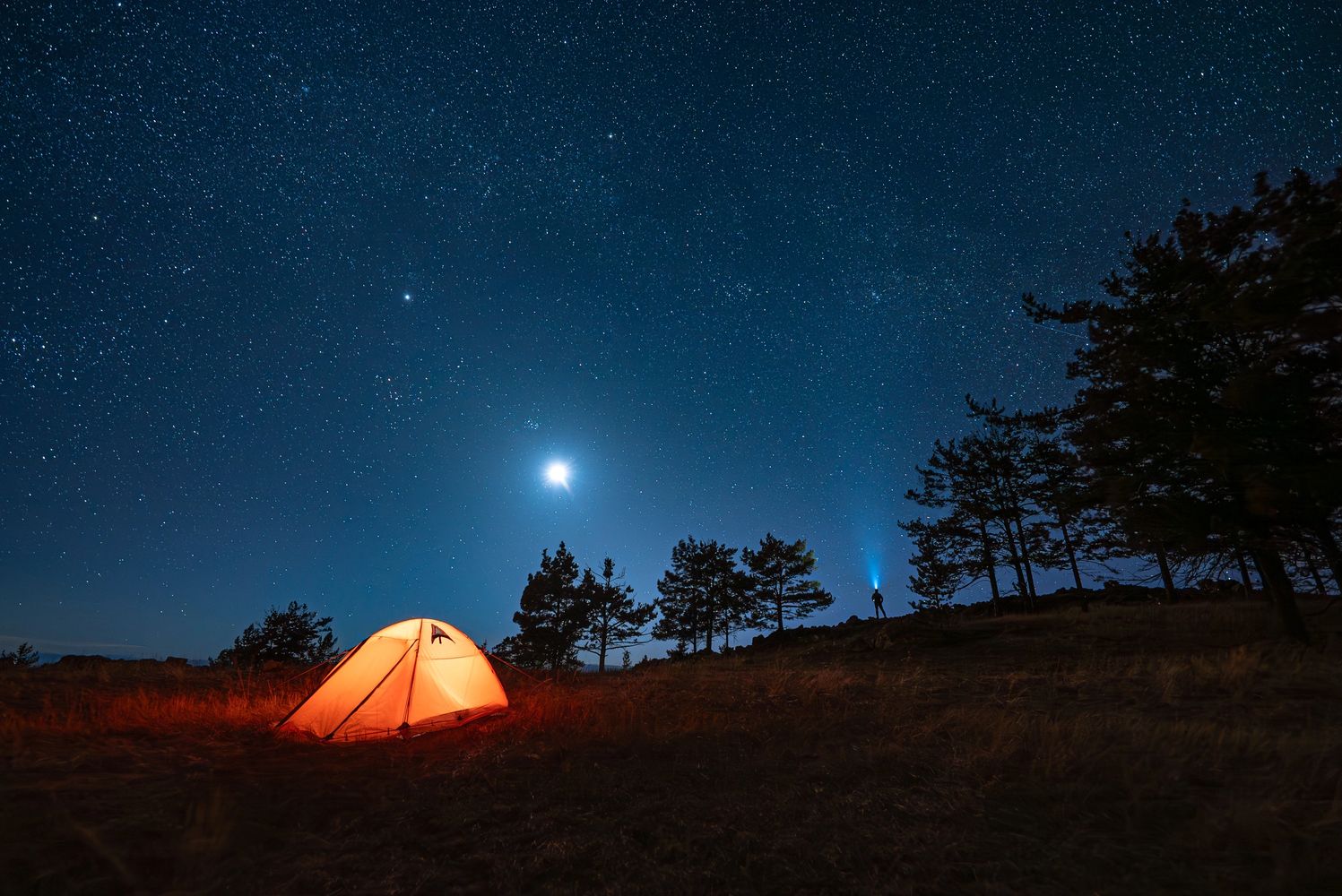 Illuminated tent under a starry night sky with a person holding a flashlight near trees.