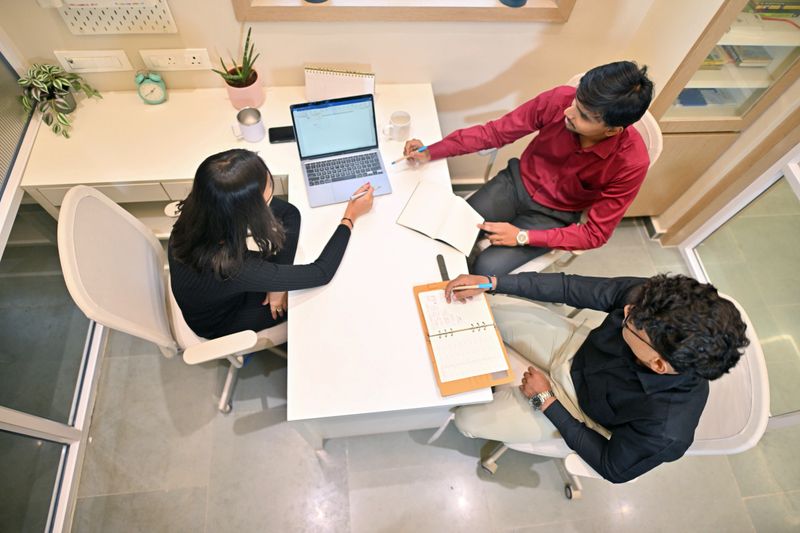 An overhead shot of a young startup team sitting together at a desk, reviewing business data on a laptop. All are engaged in discussion, pointing at graphs and taking notes. The modern workspace and collaborative energy reflect the strategic planning and teamwork typical in a startup environment.