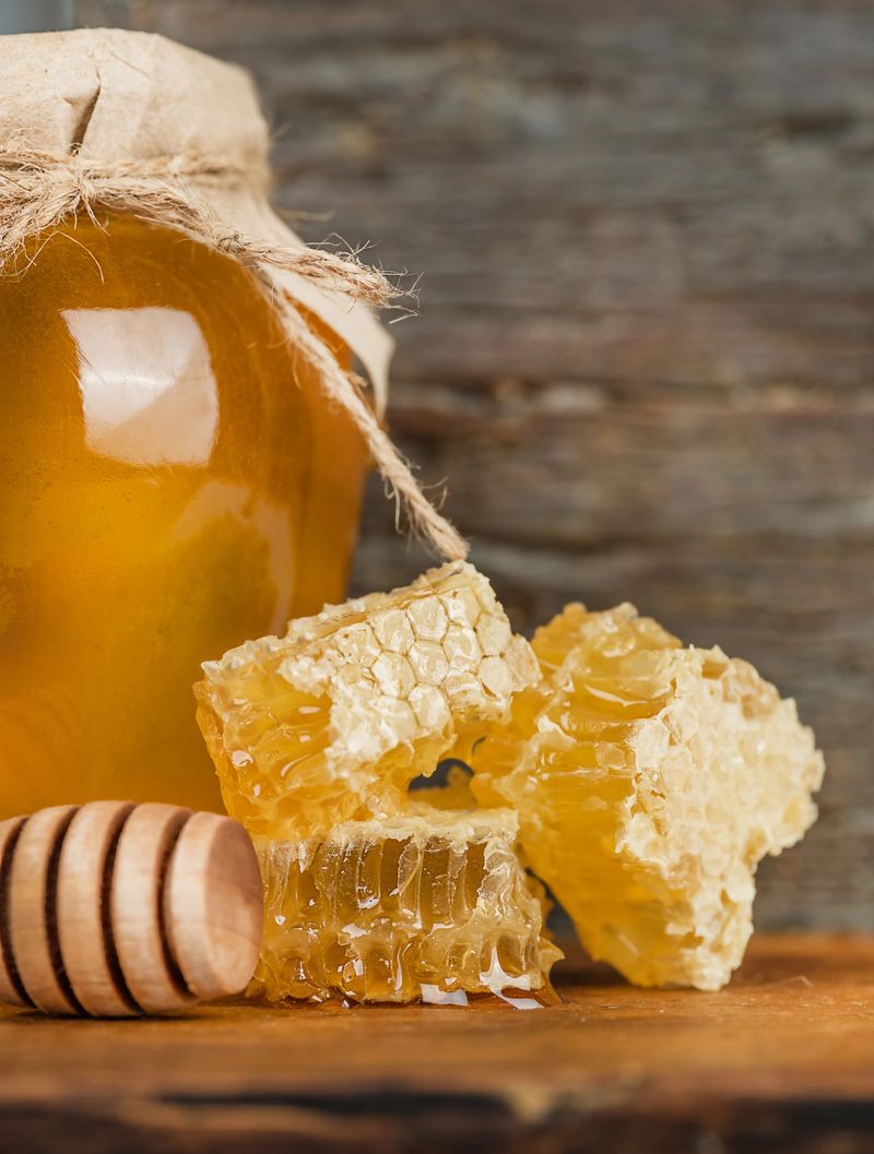 Close-up of honey jar, golden honeycomb chunks, and wooden dipper on rustic wood surface. Natural organic food concept with warm tones and healthy sweetness.