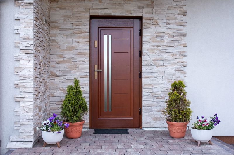 A front door detail with a stone accent wall, a brown door, covered porch, white posts and railing, and white siding. Exterior facade. External doors. Wooden doors.
