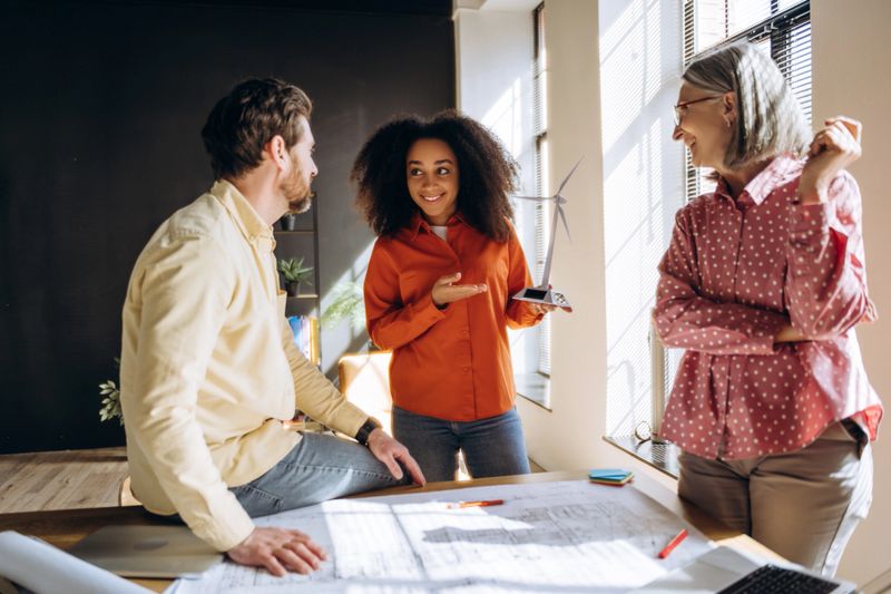 Three engineers are discussing a miniature wind turbine and solar panel model in a brightly lit office, reviewing blueprints spread out on a table, indicating a focus on renewable energy solutions. Teamwork concept