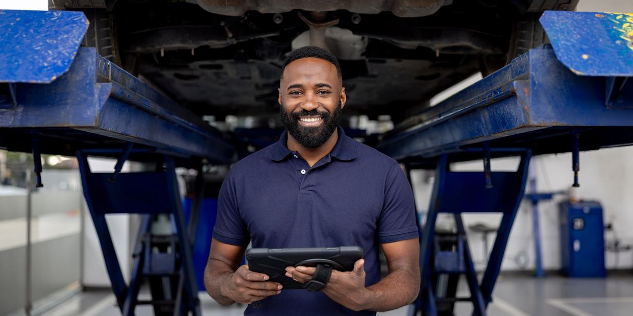 Smiling mechanic holding a tablet under a car on a lift in a garage.
