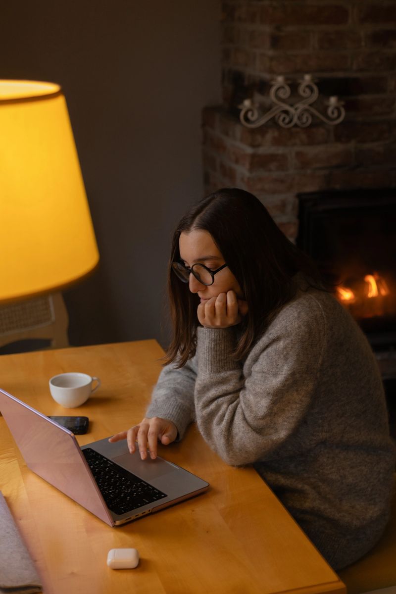 A woman works on her laptop by the fireplace, her head resting on her hand
