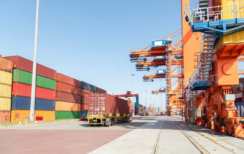 Truck carrying container and ready for leaving port terminal and quay crane standing on the background. View of Seaport operation activities, container shipping, and logistics.