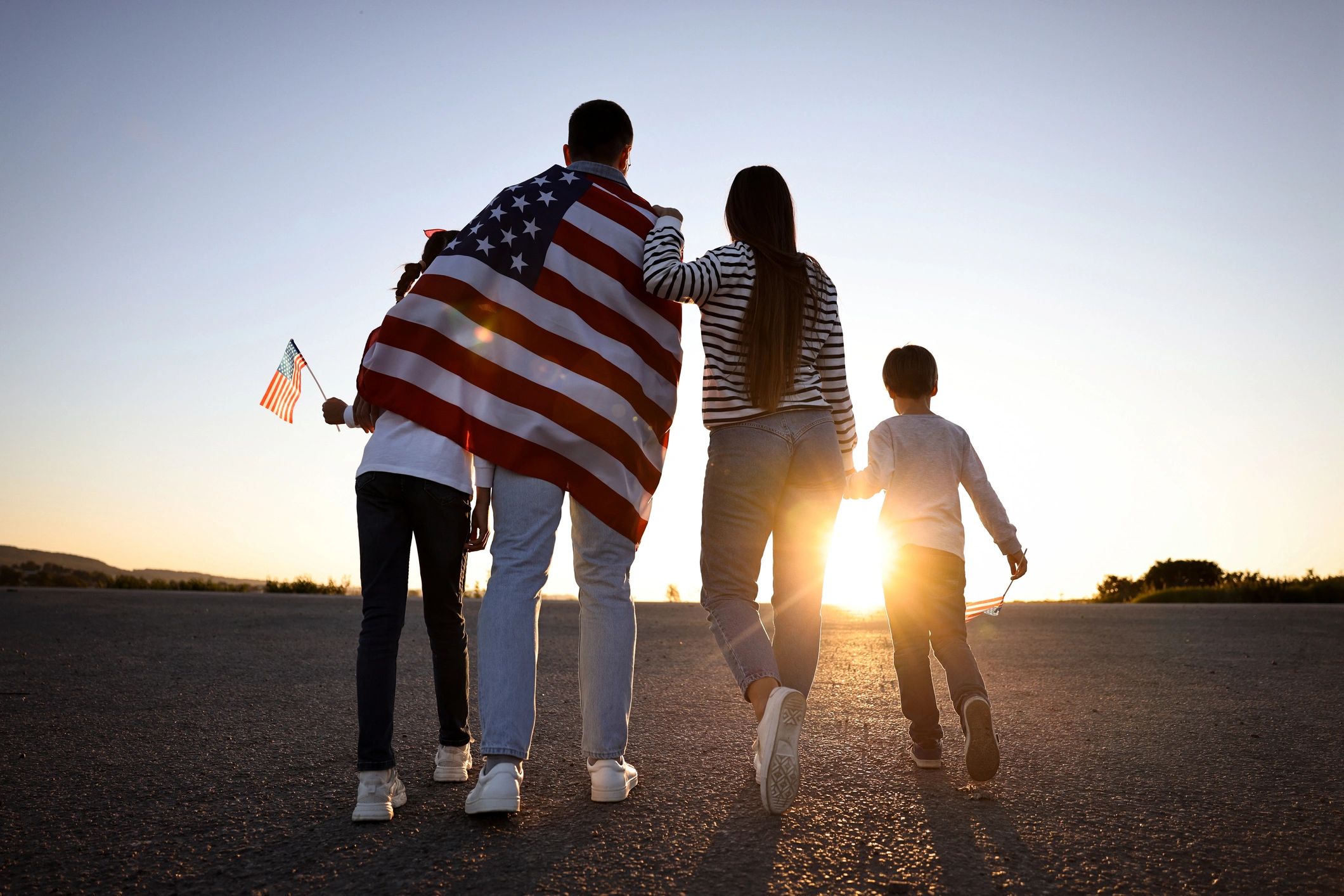 Family walking at sunset, one wrapped in an American flag, holding small flags.