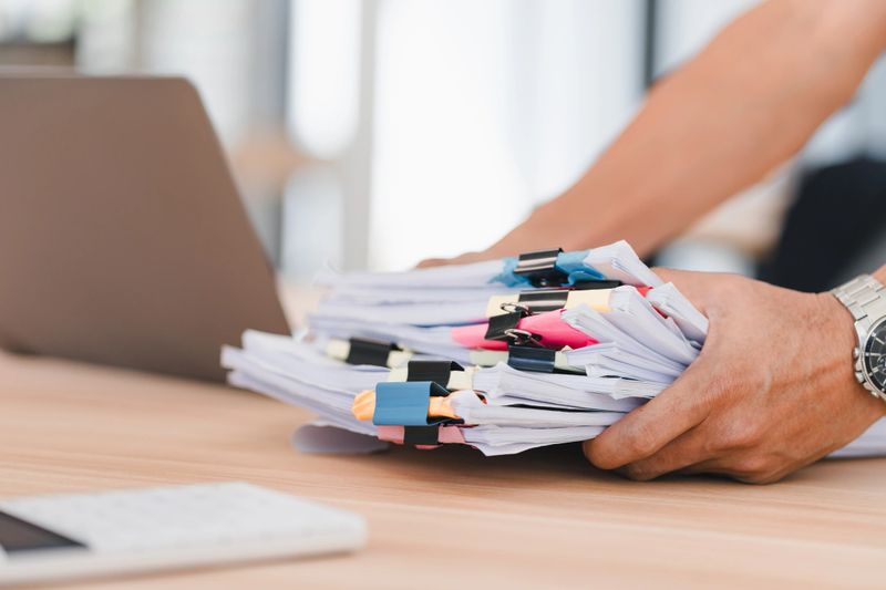 A person organizing stack of documents with colorful paper clips on wooden desk near laptop