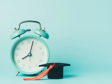 A turquoise alarm clock showing 8:02 next to a black graduation cap with an orange tassel.