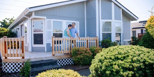 A couple stands on the porch of a blue house surrounded by greenery.