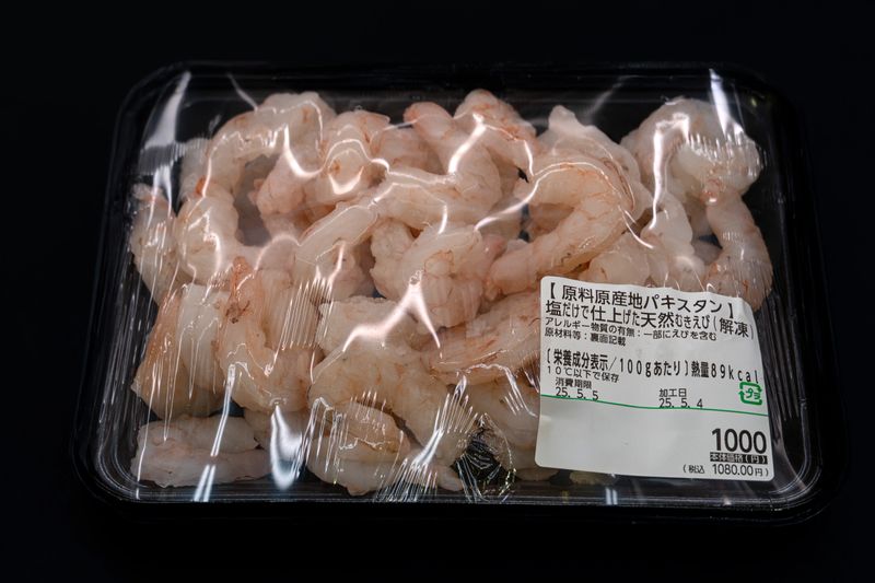 A clear, top-down shot of a plastic-wrapped package of defrosted, peeled natural shrimp imported from Pakistan. The shrimp appear to be medium-sized and have a translucent, pale pinkish-white color. Japanese labeling on the package provides details about the product's origin ("原料原産地パキスタン" - Raw material origin: Pakistan), preparation ("塩だけで仕上げた天然むき海老（解凍）" - Natural peeled shrimp finished only with salt (defrosted)), and nutritional information, including calories per 100g. The plastic packaging shows some condensation, indicating it has been defrosted.